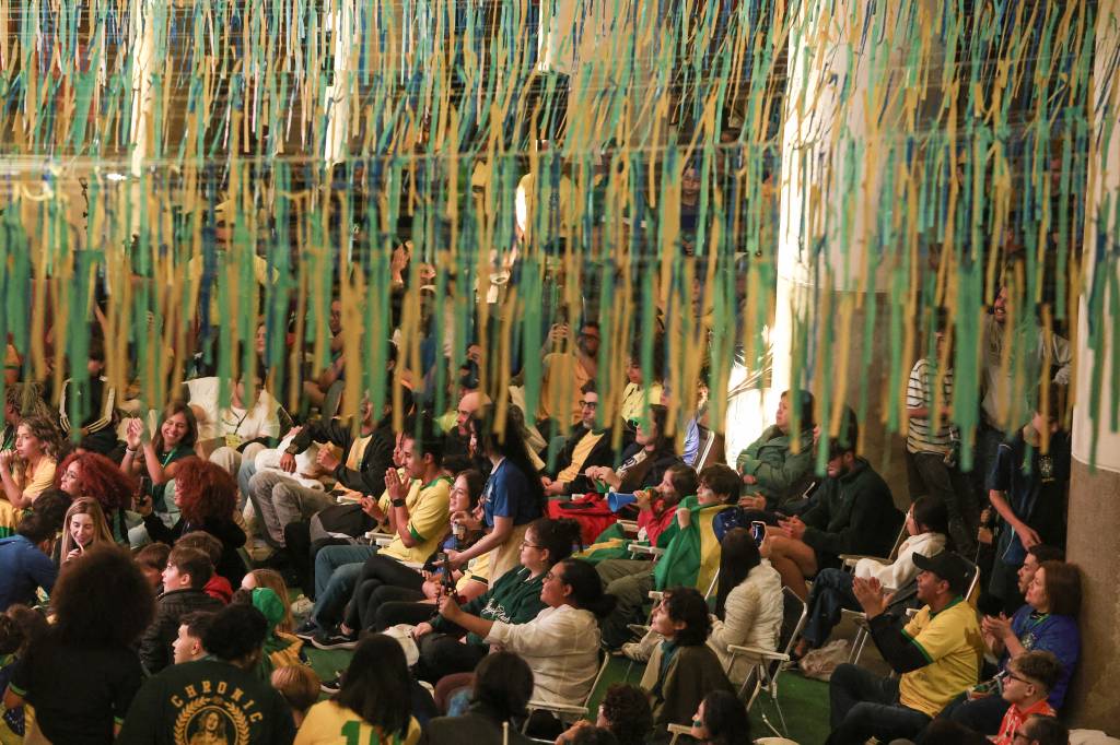 Multidão de pessoas assistindo a um evento, algumas aplaudindo, sob uma decoração de fitas verdes, azuis e amarelas penduradas no teto