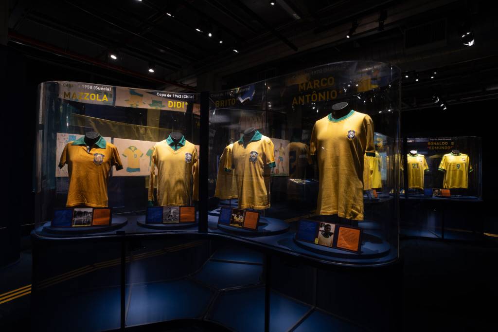 Camisas amarelas da seleção brasileira de futebol expostas em vitrines de vidro, com nomes de jogadores como Mazzola, Didi e Marco Antônio. O ambiente escuro é iluminado por focos de luz que destacam os uniformes e informações sobre as Copas de 1958 e 1962