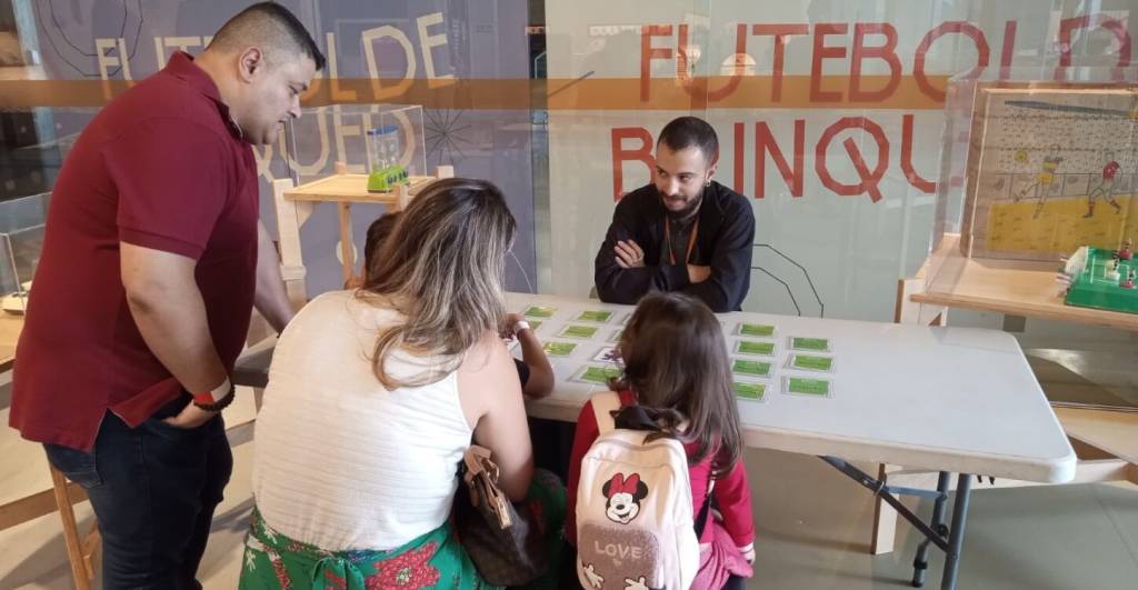 Três adultos e uma criança interagem em uma mesa com cartas verdes. Um homem de camisa polo vermelha e outro de jaqueta preta observam, enquanto uma mulher de blusa branca e uma menina de mochila da Minnie com a palavra LOVE olham para as cartas. Ao fundo, uma vitrine exibe um jogo de futebol de botão e um painel com a palavra FUTEBOL