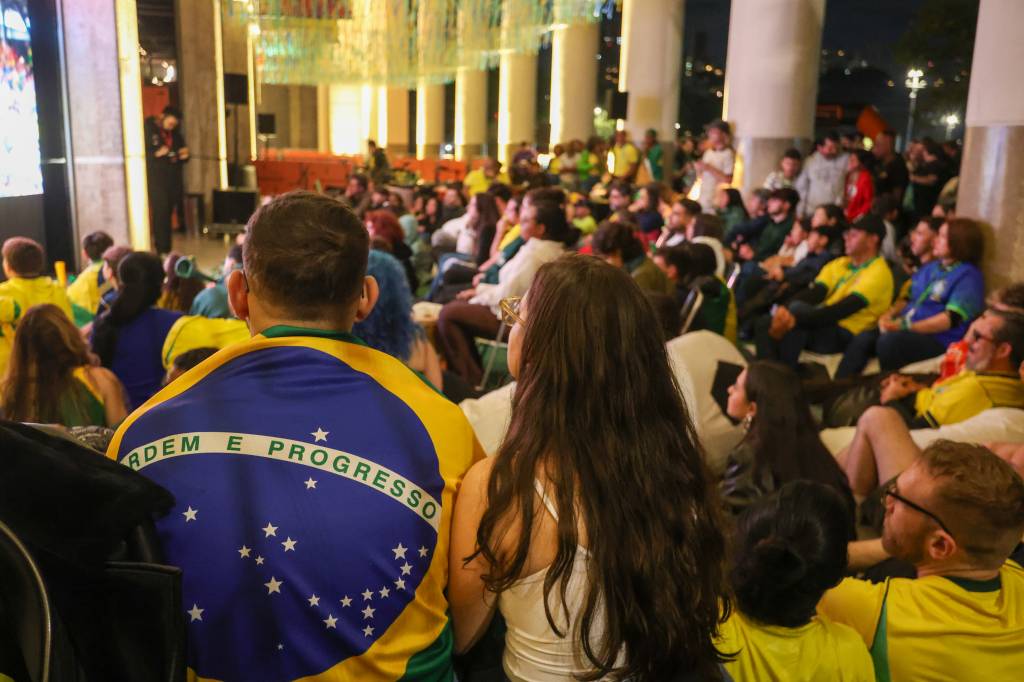 Pessoas assistem a um evento em ambiente interno, com um homem de costas vestindo uma camisa da bandeira do Brasil com ORDEM E PROGRESSO e uma mulher de cabelos longos ao lado. O público está sentado em cadeiras e pufes, alguns usando roupas com as cores da bandeira brasileira, em um espaço com iluminação indireta e decoração suspensa
