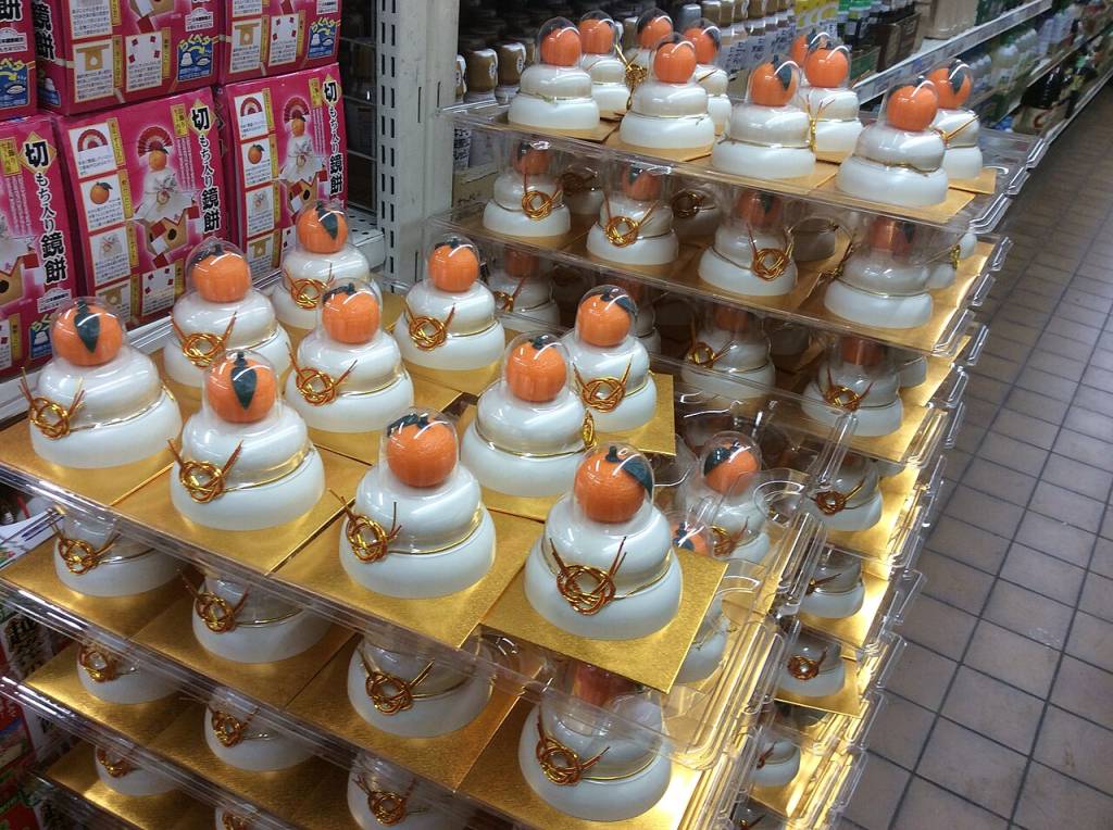 Muitos kagami mochi, bolos de arroz brancos em camadas com uma tangerina no topo e um laço dourado, expostos em prateleiras de supermercado