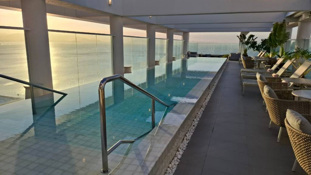 Piscina de borda infinita em um terraço com vista para o mar ao pôr do sol. A piscina tem degraus com corrimão de metal e ao lado espreguiçadeiras e cadeiras de vime com almofadas claras, sob um teto branco sustentado por pilares. O céu está dourado e o mar reflete a luz