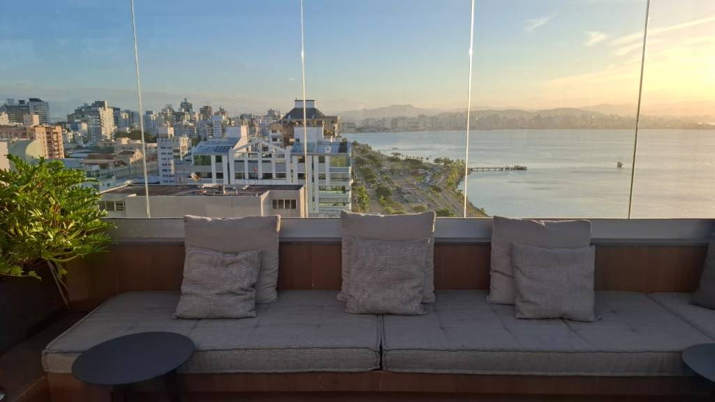 Sofás cinzas com almofadas em um terraço com vista panorâmica para uma cidade costeira, mar e montanhas ao pôr do sol