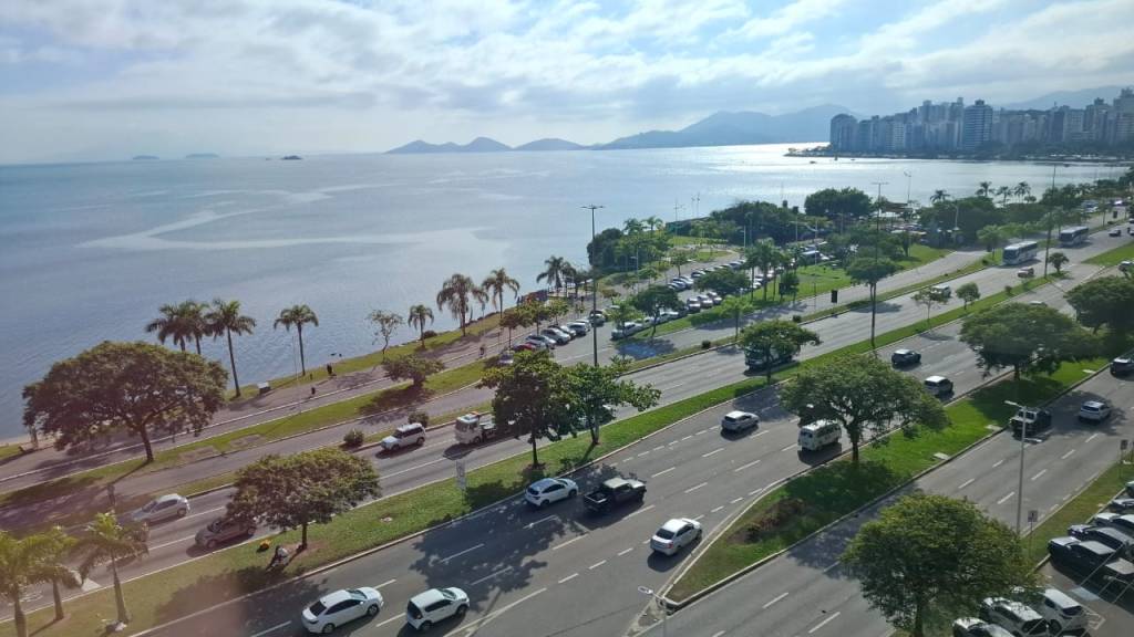 Vista aérea de uma avenida movimentada à beira-mar, com carros e ônibus circulando. À esquerda, o oceano azul-acinzentado com montanhas distantes e, à direita, prédios altos. Coqueiros e árvores verdes margeiam a via, com um céu parcialmente nublado
