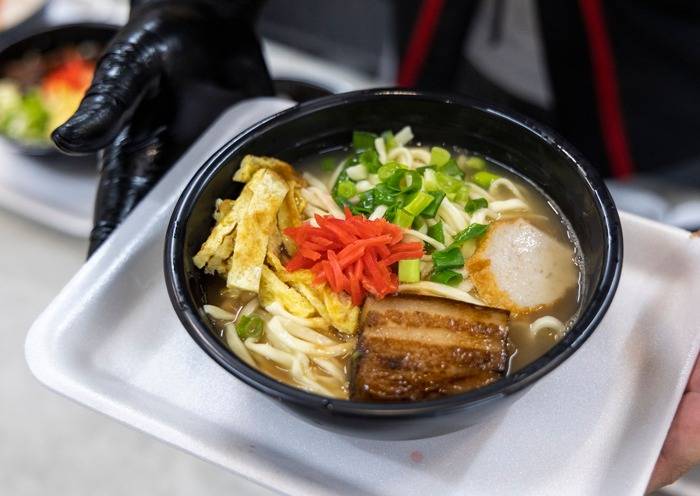 Uma pessoa com luva preta segura uma tigela de ramen com macarrão, carne de porco, omelete fatiado, cebolinha e gengibre vermelho picado, servida em uma bandeja de isopor