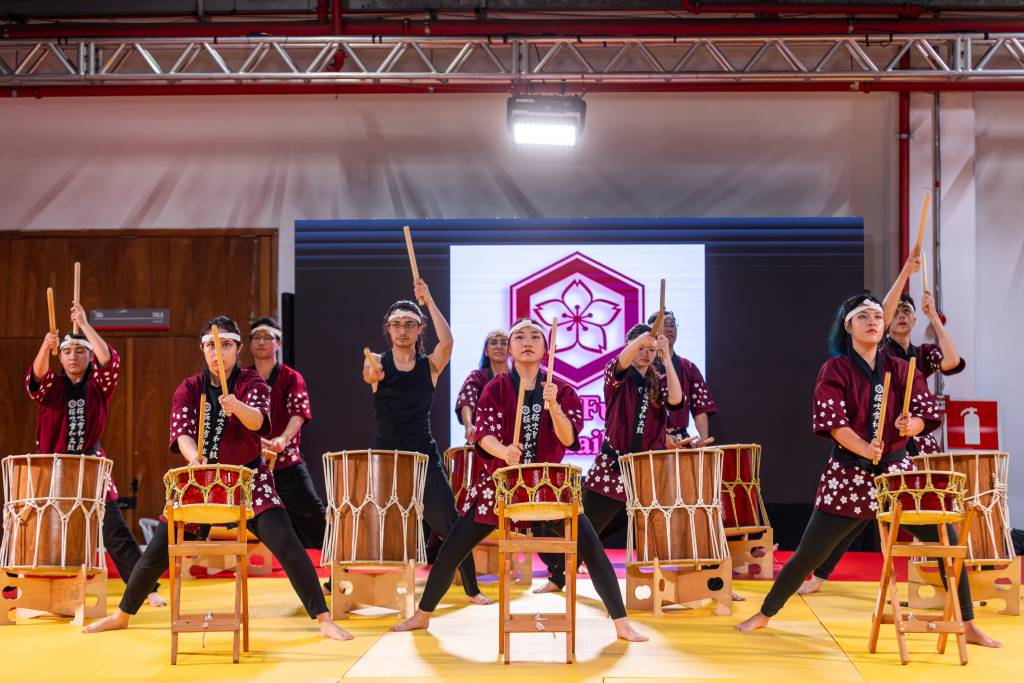 Grupo de pessoas, algumas com quimonos vermelhos e brancos, tocando tambores taiko com baquetas de madeira em um palco amarelo. Ao fundo, um telão exibe um logotipo com uma flor de cerejeira