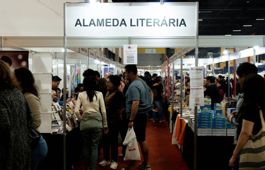 Pessoas circulam em uma feira de livros, com estandes brancos e prateleiras cheias de publicações. O letreiro principal exibe ALAMEDA LITERÁRIA em letras pretas. Um homem de camiseta azul e bermuda preta segura uma sacola branca, enquanto outros visitantes olham os livros. O chão é um carpete vermelho, e o ambiente está bem iluminado