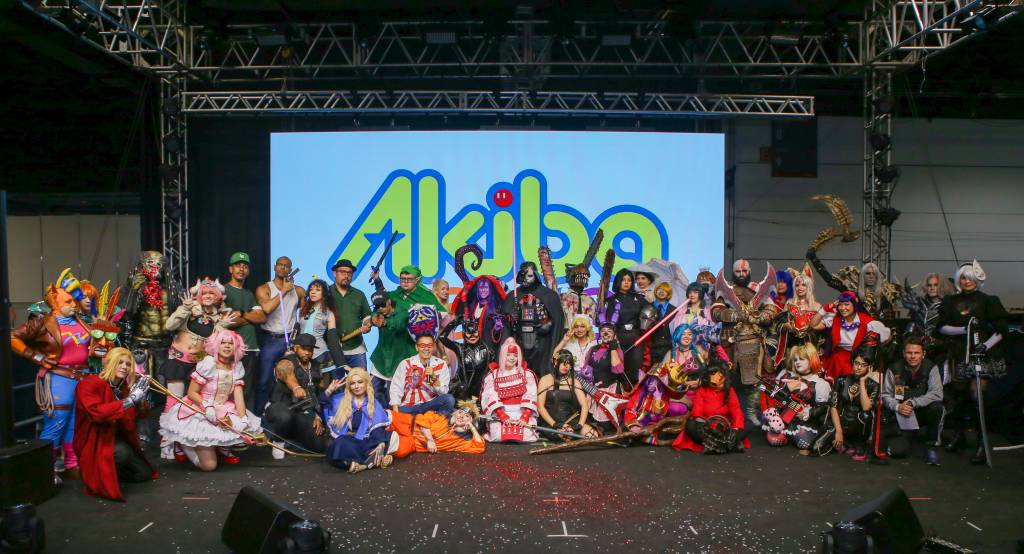 Um grande grupo de cosplayers posa para foto em palco com telão exibindo o logo Akiba. Há personagens como Darth Vader, Kratos, Predador e outros de animes e games, alguns em pé e outros agachados, com confetes espalhados pelo chão
