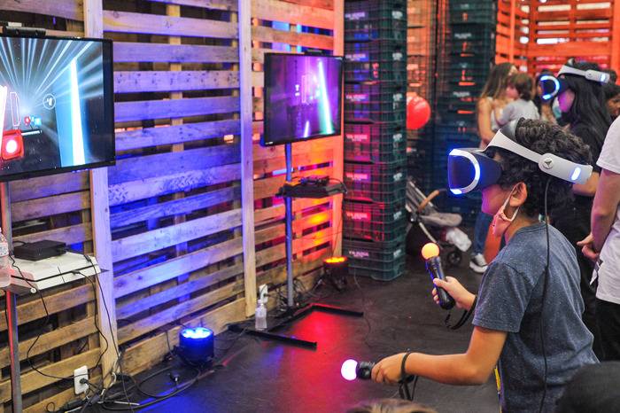 Um jovem com cabelo cacheado e camiseta azul joga videogame de realidade virtual, usando óculos e segurando controles luminosos. Ao fundo, paredes de pallets de madeira iluminadas por luzes azuis e vermelhas, e outras pessoas observando