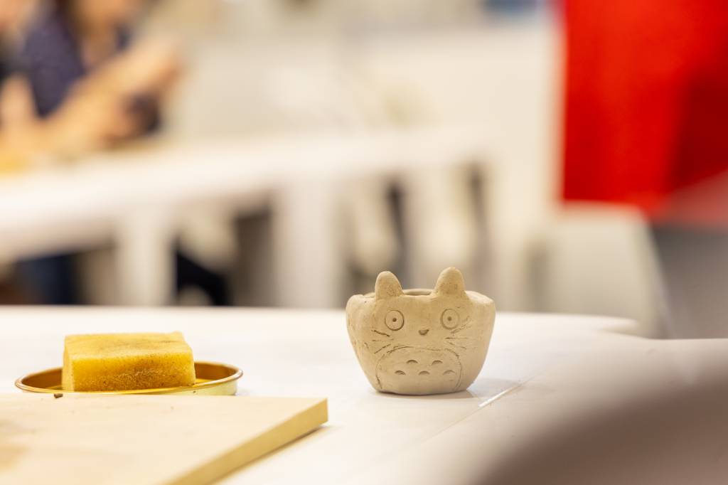 Um pequeno vaso de cerâmica bege em formato de Totoro, com orelhas pontudas e olhos grandes, está sobre uma mesa branca. À esquerda, um doce quadrado amarelo em um prato dourado. Ao fundo, pessoas desfocadas e uma mancha vermelha.