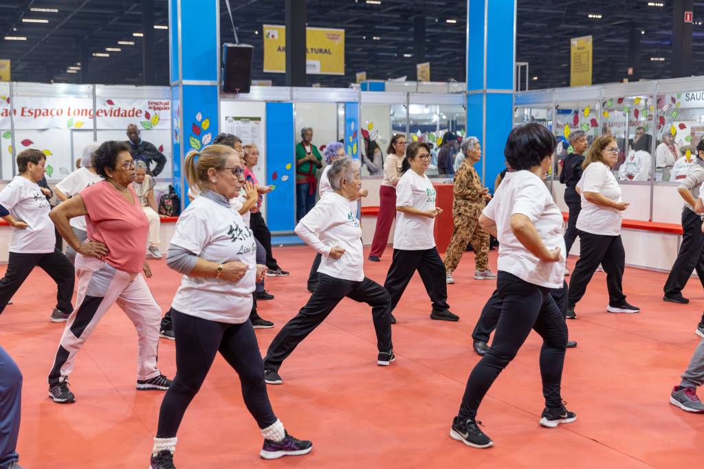 Grupo de idosos, a maioria mulheres, praticando exercícios físicos em um salão com piso vermelho. Eles usam roupas esportivas e estão em posição de alongamento, com uma perna à frente e os braços flexionados. Ao fundo, há estandes de eventos com letreiros como Espaço Longevidade e Saúde.