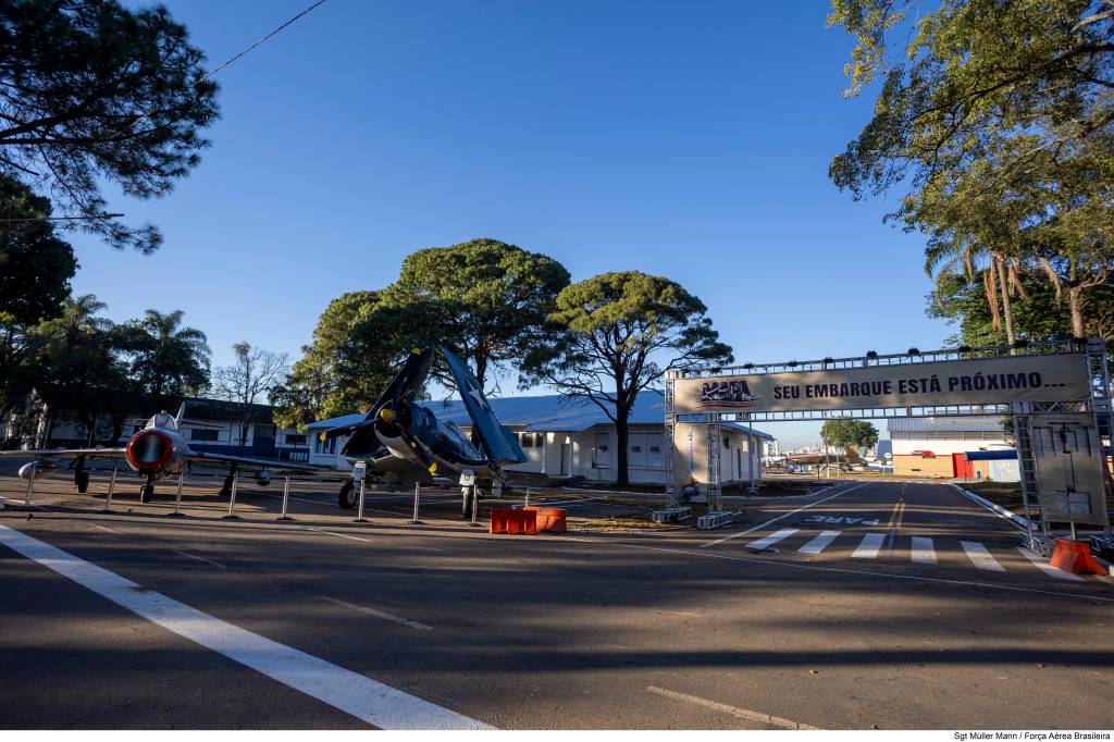 Dois aviões militares antigos em exposição, um a jato prateado e um azul com hélice, ladeiam uma estrada asfaltada. Ao fundo, um portal com a frase SEU EMBARQUE ESTÁ PRÓXIMO... e árvores frondosas sob céu azul claro