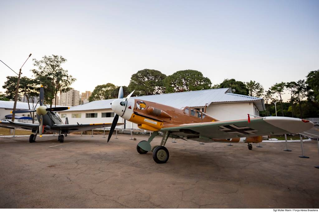 Dois aviões de guerra antigos em exposição, um cinza à esquerda e um marrom-amarelado à direita, com hélices e asas marcadas com cruzes pretas, em um pátio de concreto com prédios brancos e árvores ao fundo
