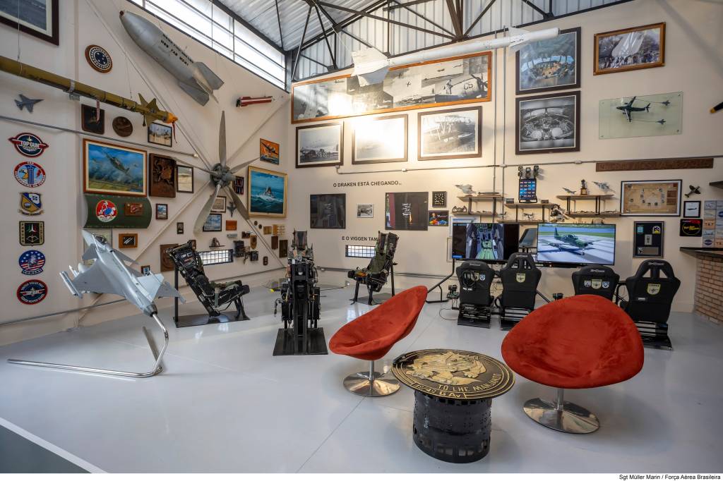 Ambiente interno de um museu ou sala temática de aviação, com paredes brancas repletas de quadros, maquetes de aviões e mísseis pendurados. No centro, uma hélice de avião grande. Há cadeiras de simulação de voo e poltronas vermelhas, com telas exibindo imagens de aeronaves. O chão é cinza claro