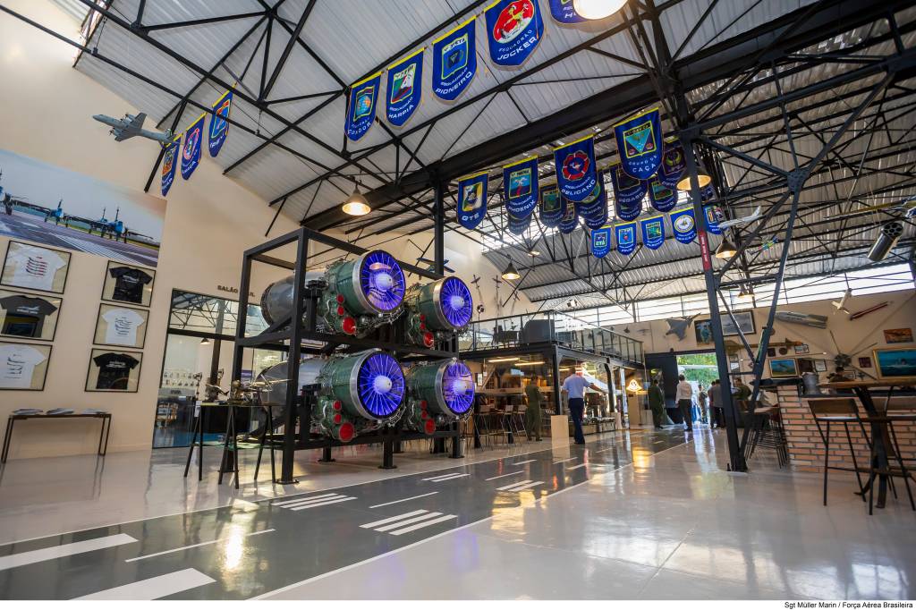 Interior de um museu ou hangar com quatro motores de avião em exposição, dois em cada nível de uma estrutura preta. Os motores têm turbinas azuis iluminadas. O chão cinza simula uma pista de aeroporto. Bandeiras azuis com emblemas pendem do teto. Pessoas caminham ao fundo, e camisetas emolduradas decoram uma parede. Um modelo de avião está suspenso no alto