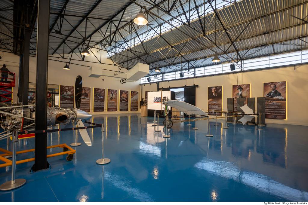 Interior de um museu de aviação com piso azul brilhante. À esquerda, um motor de avião com hélice e um homem em uma plataforma elevatória. Ao fundo, painéis informativos com fotos de aviadores e um modelo de aeronave antiga. O teto é de metal corrugado com vigas expostas e luminárias penduradas