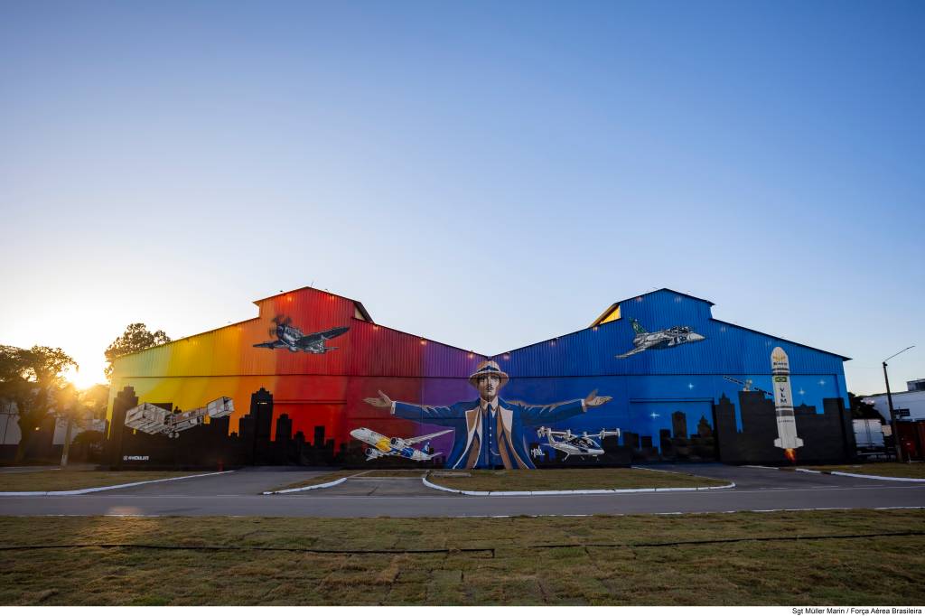 Mural colorido em dois galpões, com um homem de chapéu e terno azul de braços abertos ao centro. À esquerda, um avião antigo e um moderno sobre um fundo vermelho e laranja. À direita, um caça e um foguete sobre um fundo azul. O sol brilha no horizonte esquerdo