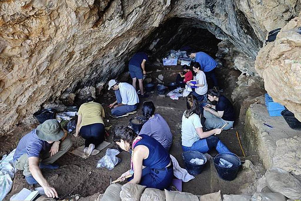 Arqueólogos, homens e mulheres, escavam cuidadosamente o chão de uma caverna rochosa, usando ferramentas pequenas e baldes, em um sítio arqueológico