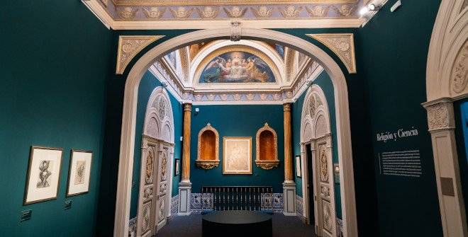 Um salão de museu com paredes verde-escuras, teto abobadado decorado e um arco central que revela outro ambiente. No centro, uma pintura emoldurada entre dois nichos dourados. À esquerda, duas gravuras emolduradas. À direita, um texto na parede com o título Religión y Ciencia