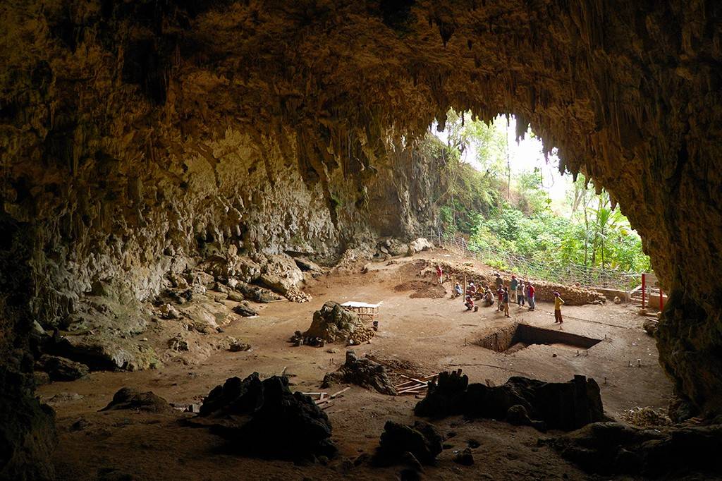 Interior de uma caverna ampla e escura, com teto e paredes rochosas irregulares, cheias de estalactites. No fundo, um grupo de arqueólogos trabalha em escavações, com a luz do dia e vegetação exuberante visíveis na entrada da caverna