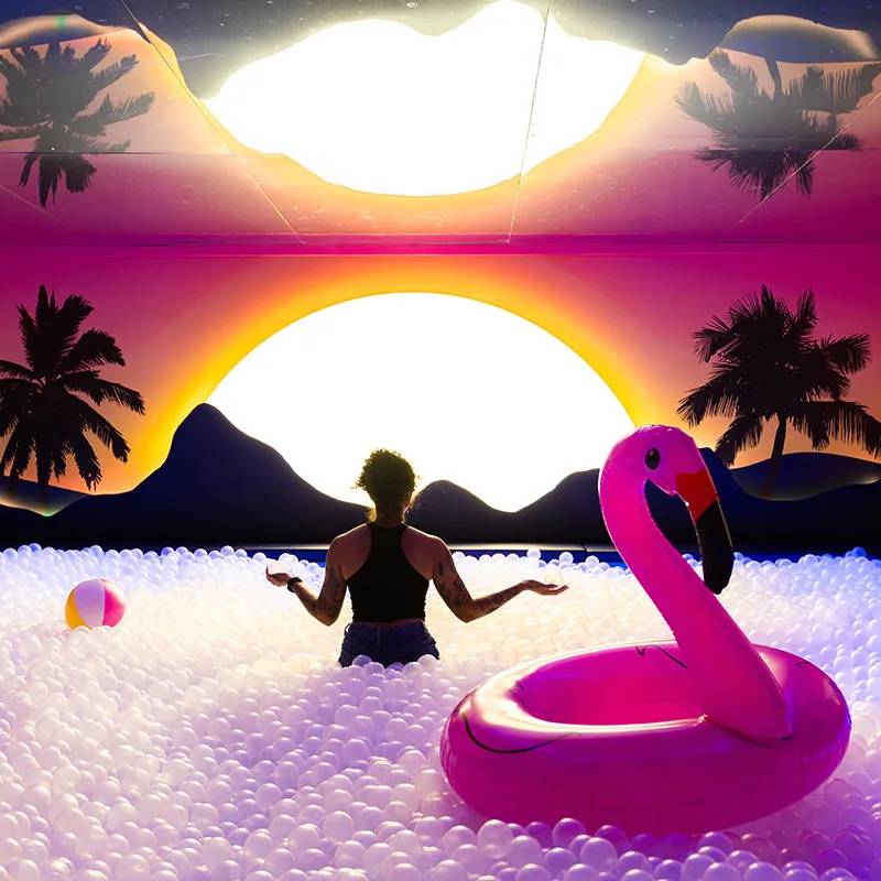 Mulher de costas, com top preto e shorts jeans, imersa em piscina de bolinhas brancas, braços abertos. Ao fundo, um pôr do sol vibrante com palmeiras e montanhas escuras. À direita, uma boia de flamingo rosa e à esquerda, uma bola de praia listrada. A cena reflete-se no teto