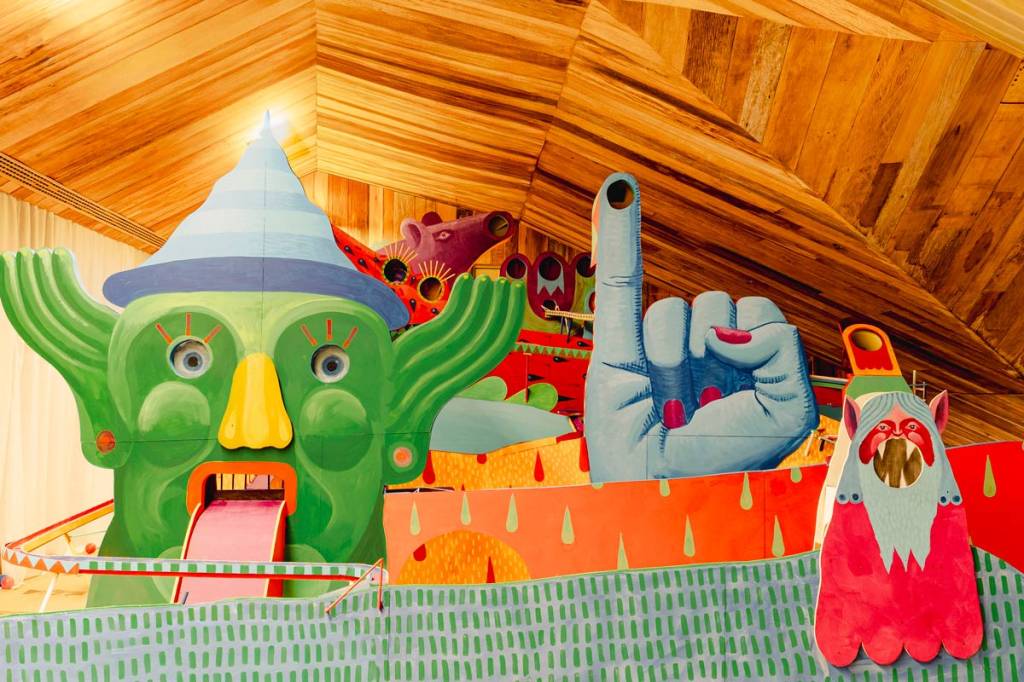 Instalação artística colorida com figuras de desenho animado: um monstro verde de chapéu azul, uma mão azul com dedo indicador levantado e um ser rosa com barba branca, sobre um fundo de madeira