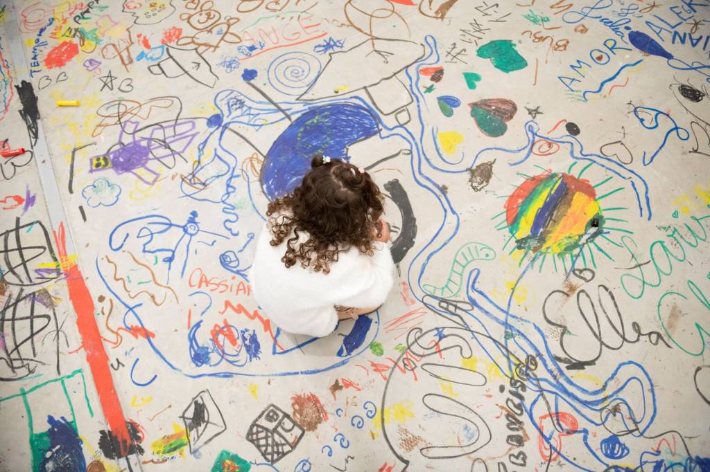 Criança de cabelos cacheados, vista de cima, sentada no chão coberto por desenhos coloridos de giz de cera, incluindo um sol arco-íris e a palavra AMOR