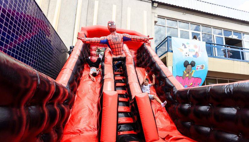 Crianças brincam em um pula-pula inflável gigante vermelho e preto, com um Homem-Aranha no topo. Um menino desce de cabeça para baixo à esquerda, outro sobe as escadas centrais e um terceiro desce sentado à direita, com os braços para cima. Ao fundo, um prédio com janelas e uma faixa azul com a frase Férias Mágicas