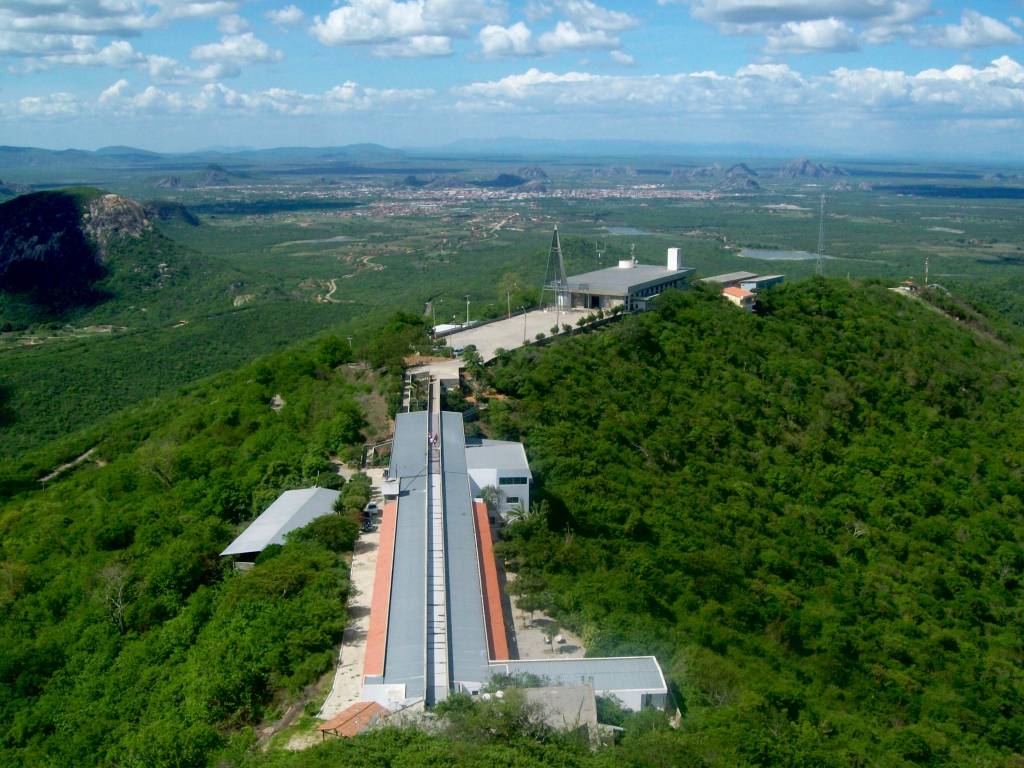 Vista aérea de um complexo de edifícios modernos no topo de uma colina verdejante, com uma longa passarela coberta cinza e telhado de terracota. Ao fundo, uma cidade se estende por um vale, cercada por montanhas e vegetação densa sob um céu azul com nuvens brancas
