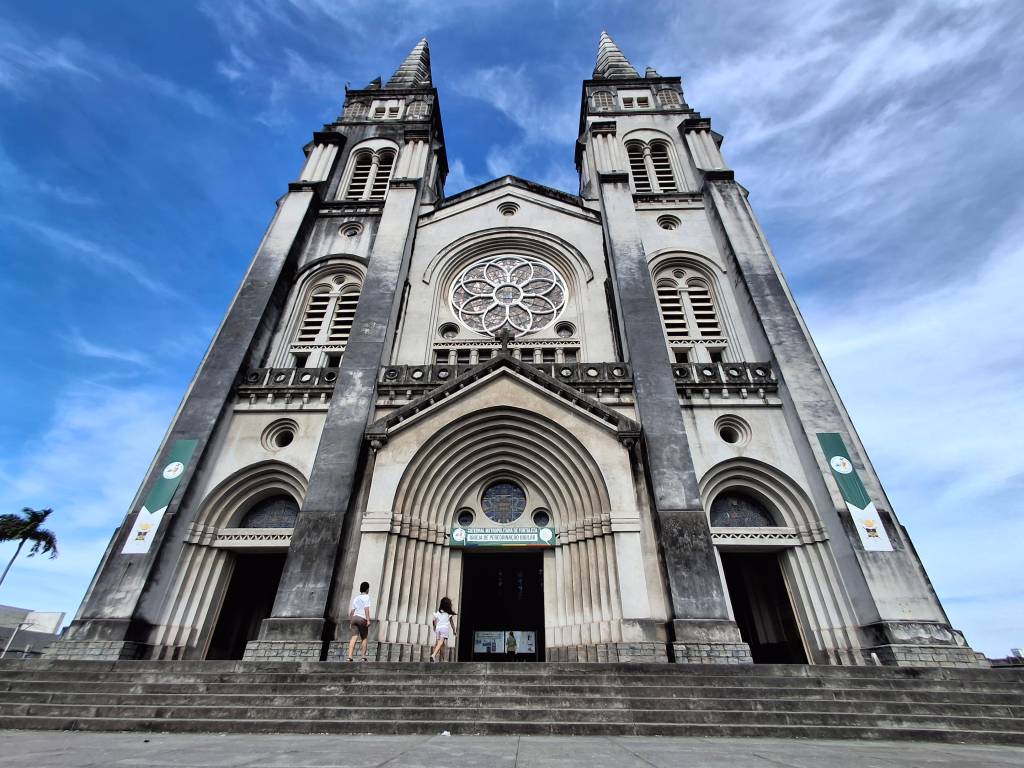 Fachada imponente da Basílica Santuário de Nazaré, em Belém, com duas torres pontiagudas e um grande rosáceo central. Dois visitantes sobem os degraus em direção à entrada principal, sob um céu azul com nuvens esparsas. Faixas verdes com símbolos religiosos adornam as laterais da construção cinza