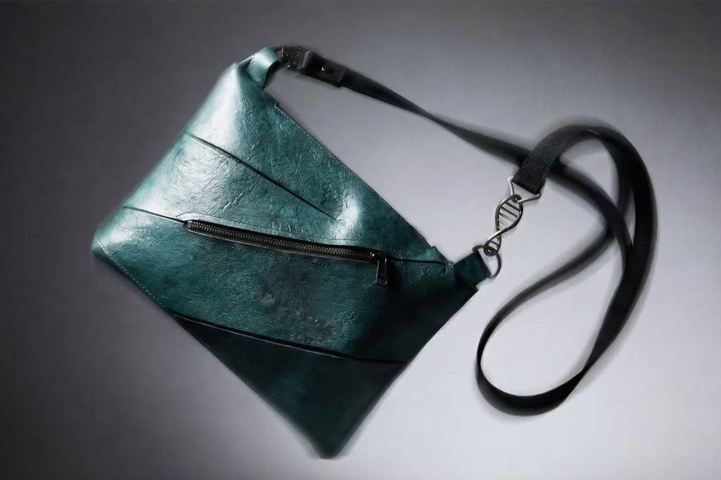Bolsa transversal verde-escura com textura enrugada, zíper frontal preto e alça preta com um chaveiro em formato de dupla hélice de DNA