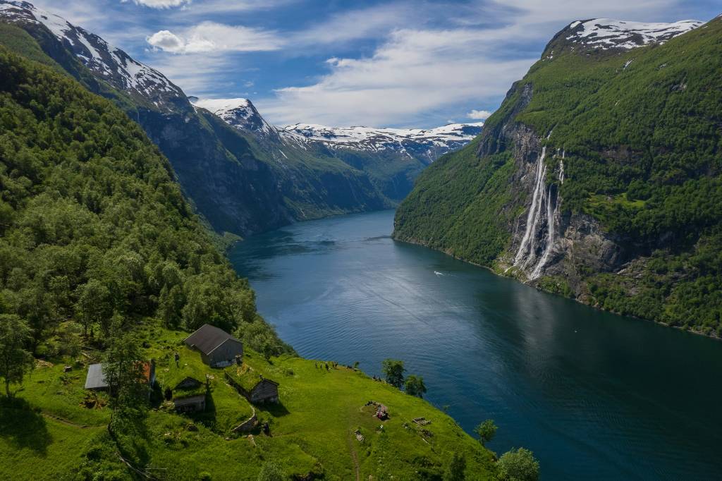Fiorde norueguês com águas azuis escuras, cercado por montanhas íngremes cobertas de florestas verdes e picos nevados. Uma cachoeira desce pela montanha à direita, e pequenas casas rústicas com telhados de grama se aninham na encosta esquerda. O céu tem nuvens brancas esparsas