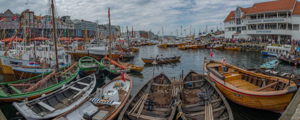 Porto movimentado com diversos barcos de madeira e veleiros ancorados, alguns com bandeiras norueguesas. Pessoas caminham nas docas e em um prédio branco com letreiros RESTAURANT - MIKROBRYGGERI e ESPRESSOBAR. O céu está nublado