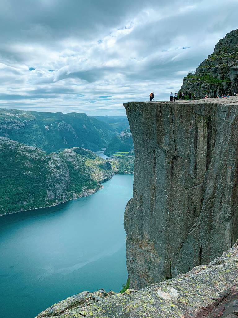 Pessoas observam a paisagem do topo do Preikestolen, um penhasco rochoso com vista para um fiorde azul-esverdeado e montanhas verdes sob um céu nublado