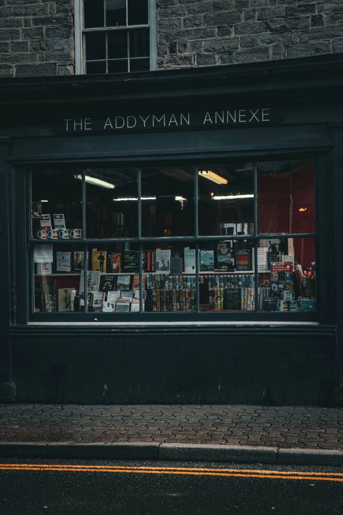 Fachada de uma livraria escura com a placa THE ADDYMAN ANNEXE em letras brancas. A vitrine exibe diversos livros, canecas e outros itens. Abaixo, uma calçada de paralelepípedos e uma rua com duas faixas amarelas
