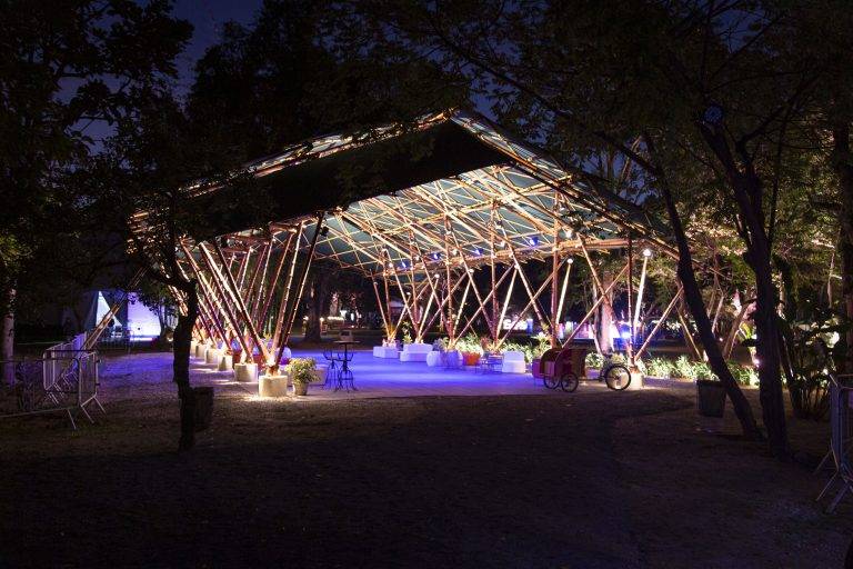 Estrutura de bambu iluminada com luzes quentes e teto escuro, em um ambiente noturno com árvores e iluminação azul no chão, criando uma atmosfera festiva