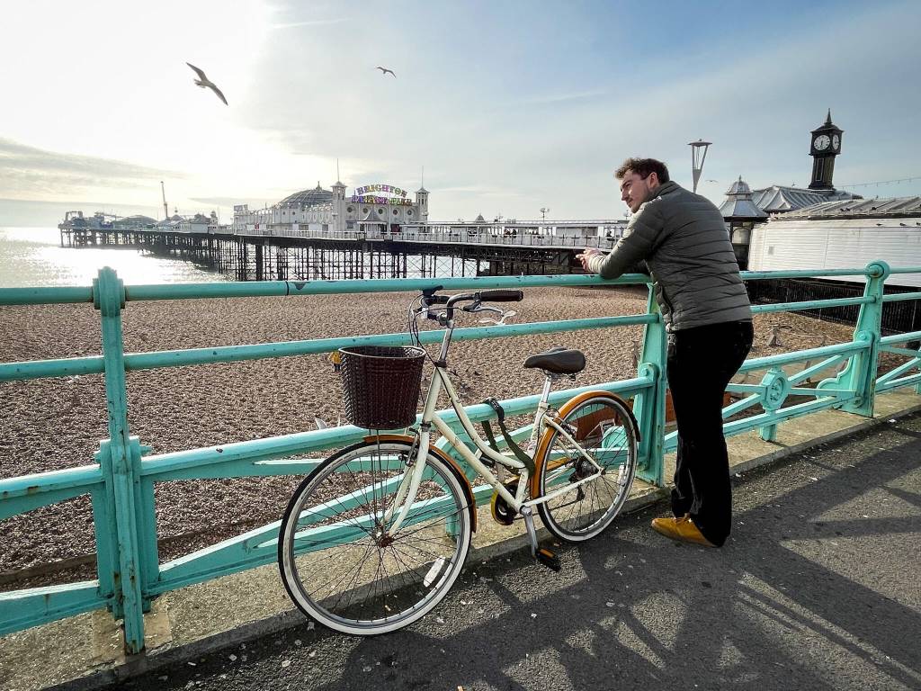 Homem de jaqueta cinza e calça preta, apoiado em uma grade verde-água, observa o mar e o píer de Brighton ao fundo. Uma bicicleta creme com cesta marrom está estacionada ao lado dele. O céu está claro, com gaivotas voando e a praia de pedras visível.