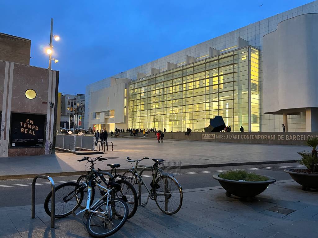 Fachada do MACBA, Museu de Arte Contemporânea de Barcelona, iluminada em tom amarelo-claro ao entardecer, com pessoas na praça e bicicletas estacionadas em primeiro plano. Foto sob licença CC BY 2.0