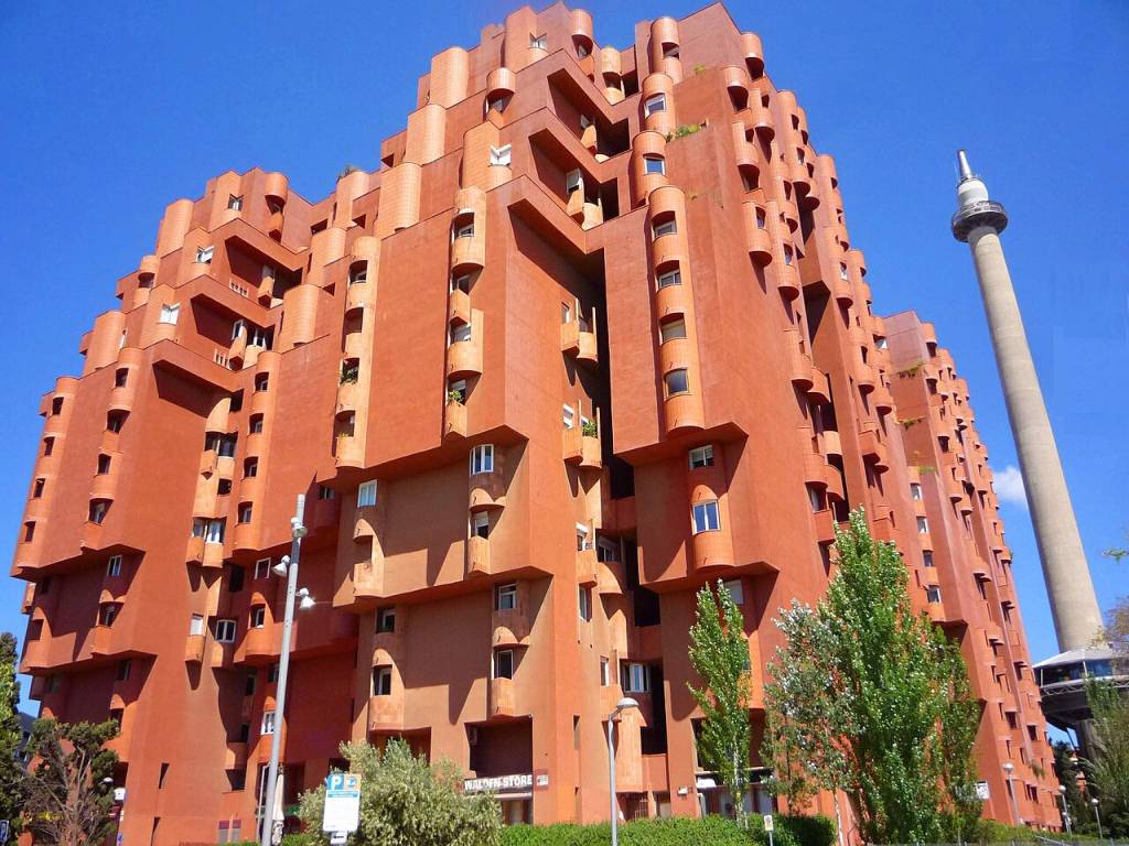 Edifício residencial de arquitetura brutalista em tons de terracota, com varandas cilíndricas e janelas quadradas, sob céu azul. À direita, uma torre de telecomunicações cinza se eleva. Árvores verdes em primeiro plano. Foto em domínio público sob licença CC0