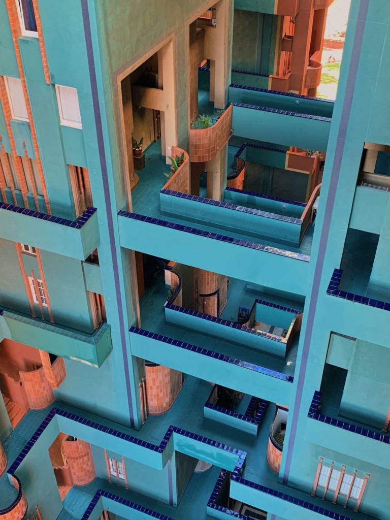 Vista aérea de um edifício com arquitetura labiríntica, paredes em tons de azul-petróleo e detalhes em tijolos alaranjados. Corredores e varandas com guarda-corpos azuis escuros se entrelaçam em vários níveis, criando um padrão complexo. Algumas plantas em vasos decoram os espaços, e janelas brancas pontuam as fachadas. A luz do sol ilumina partes da estrutura, realçando as cores vibrantes. Foto sob licença CC BY-SA 4.0