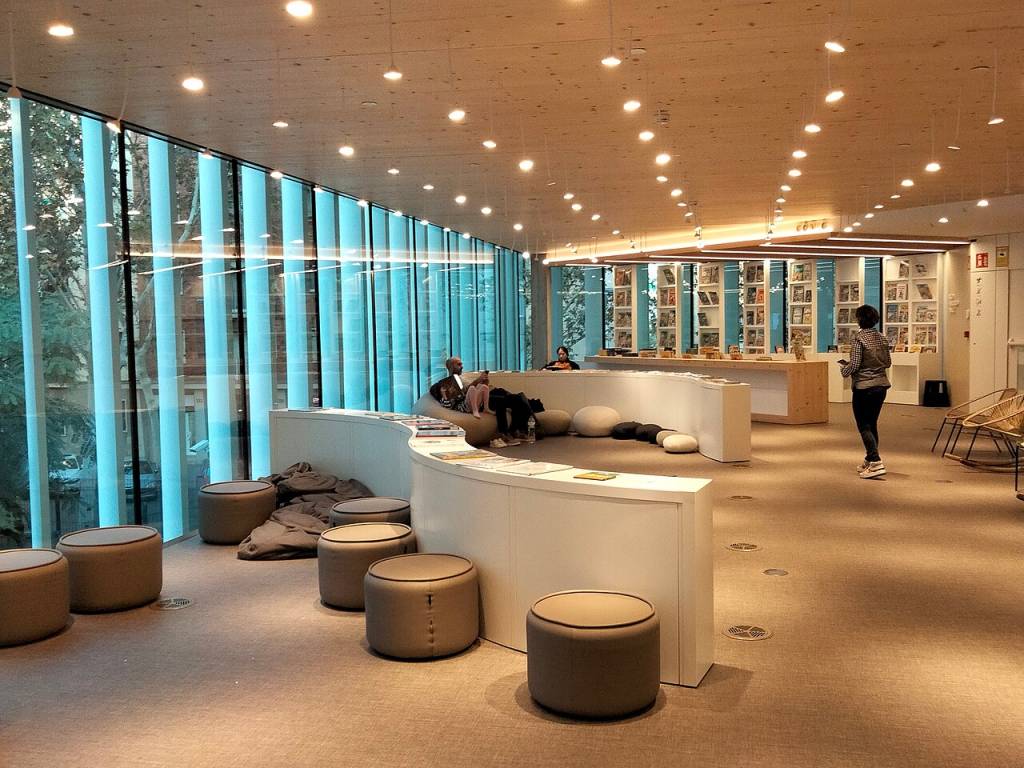 Interior de uma biblioteca moderna com teto de madeira e iluminação pontual. À esquerda, uma parede de vidro com persianas azuis verticais e vista para árvores. No centro, balcões curvos brancos com pessoas sentadas em pufes e almofadas. Uma mulher de camisa xadrez caminha à direita. Foto sob licença CC BY 4.0