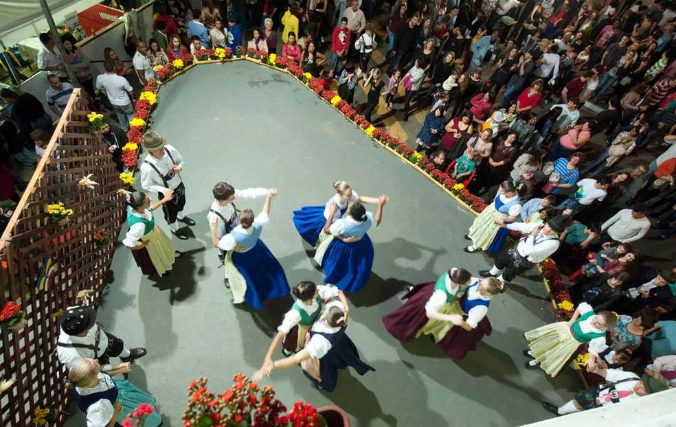 Vista aérea de um grupo de dançarinos folclóricos alemães, com homens e mulheres em trajes típicos, apresentando-se em um palco cercado por uma plateia animada. O palco é delimitado por vasos de flores coloridas, e a iluminação destaca a performance.