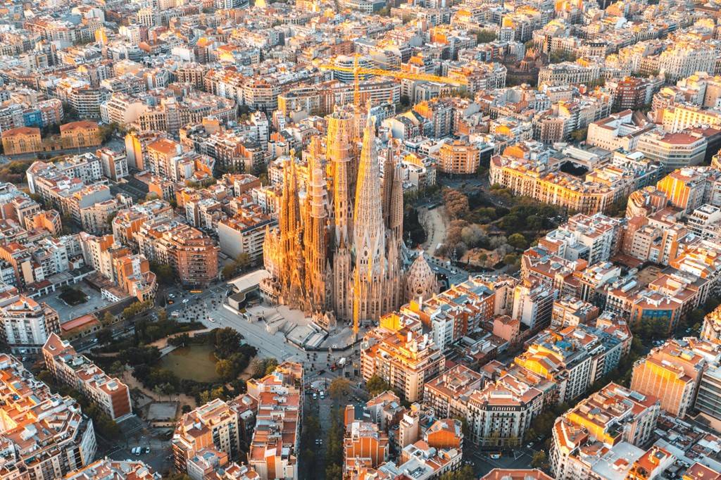 SI_489_Sagrada_Família_4 Vista aérea da Sagrada Família em Barcelona, com suas torres góticas iluminadas pelo sol poente, cercada por quarteirões de edifícios residenciais e comerciais de cor clara, com telhados alaranjados e ruas movimentadas. Há um parque com lago à esquerda da basílica e guindastes de construção visíveis ao fundo, indicando a expansão urbana