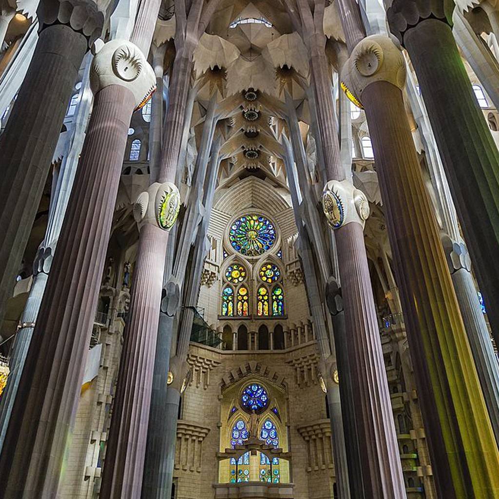 SI_489_Sagrada_Família_7 Interior da Basílica da Sagrada Família, com colunas altas e ramificadas que se assemelham a árvores, sustentando um teto abobadado. Há vitrais coloridos em formato circular e retangular, iluminando o ambiente com tons vibrantes de azul, verde, amarelo e vermelho. A arquitetura gótica modernista é grandiosa e detalhada