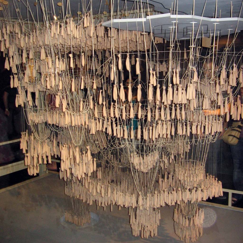 SI_489_Sagrada_Família_8 Instalação artística com centenas de pequenos sacos de tecido bege pendurados por fios finos, formando uma estrutura densa e orgânica que se estende do teto ao chão, com pessoas observando ao fundo