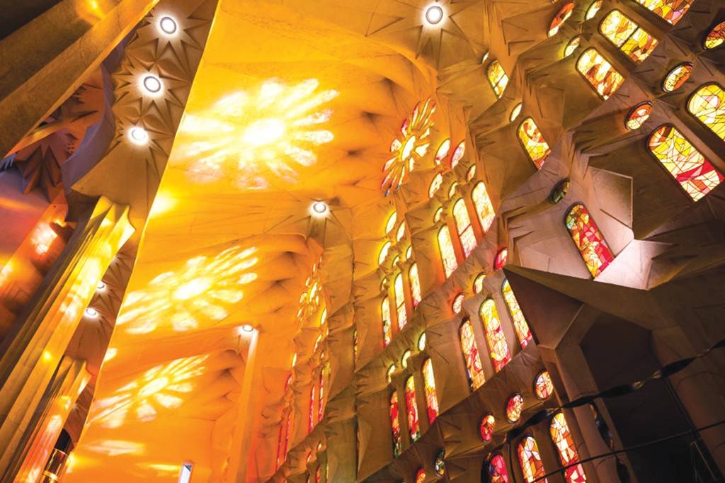 SI_489_Sagrada_Família_6 Interior de uma igreja com teto alto e abobadado, iluminado por luzes circulares e projeções de luz em formato de estrela. À direita, uma parede com múltiplos vitrais coloridos em tons de vermelho, laranja e amarelo, criando um ambiente quente e vibrante