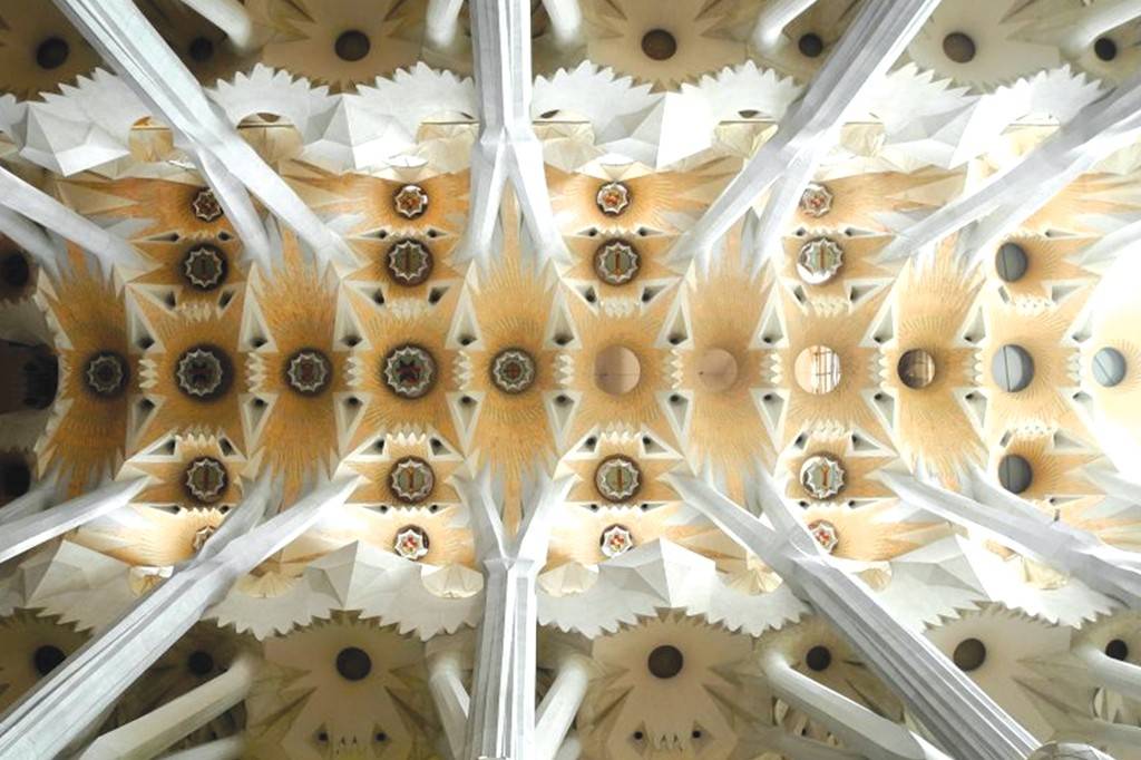 SI_489_Sagrada_Família_11 Teto de uma igreja com colunas brancas que se ramificam, sustentando painéis geométricos em tons de bege e branco, com detalhes circulares e estrelados