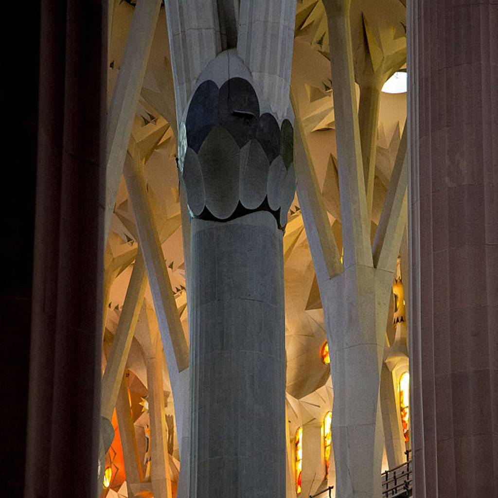 SI_489_Sagrada_Família_10 Interior da Sagrada Família com colunas ramificadas que se assemelham a árvores, iluminadas por luzes quentes e vitrais coloridos. Uma coluna central cinza tem um capitel decorado com formas ovais escuras, e outras colunas claras se estendem para o teto, criando um ambiente grandioso e complexo