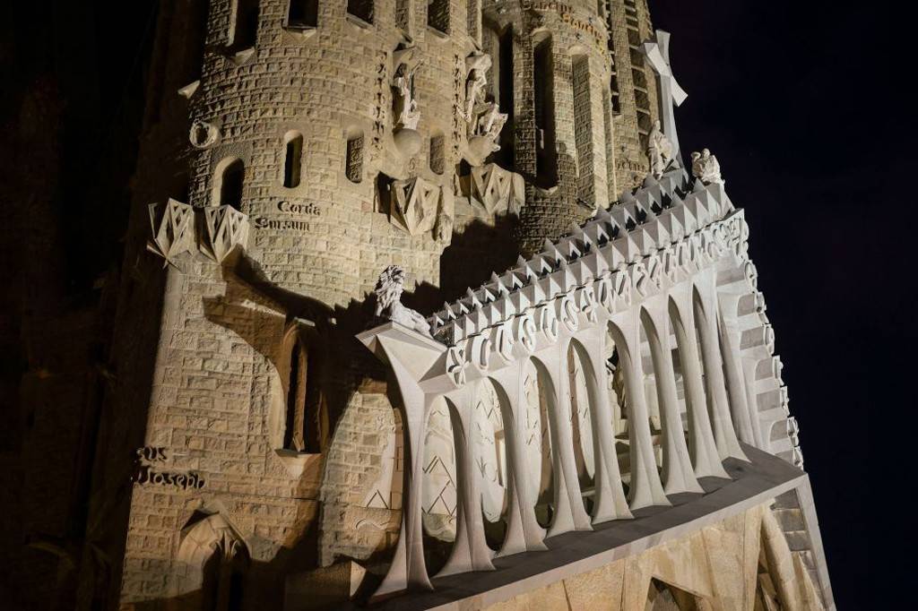 SI_489_Sagrada_Família_9 Fachada da Sagrada Família à noite, com iluminação destacando a torre de pedra e uma estrutura branca com arcos e pináculos triangulares. Esculturas e inscrições latinas como Corda Surcum e Sus Joseph são visíveis, contra um céu escuro