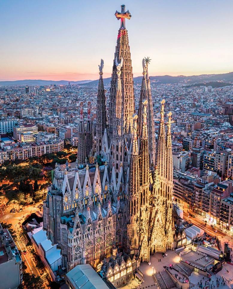 SI_489_Sagrada_Família_1 Vista aérea da Basílica da Sagrada Família em Barcelona, Espanha, ao entardecer. A arquitetura gótica da igreja é iluminada por luzes quentes, destacando suas torres pontiagudas e detalhes intrincados. Ao fundo, a cidade se estende com edifícios e ruas iluminadas, sob um céu em tons de azul e rosa.